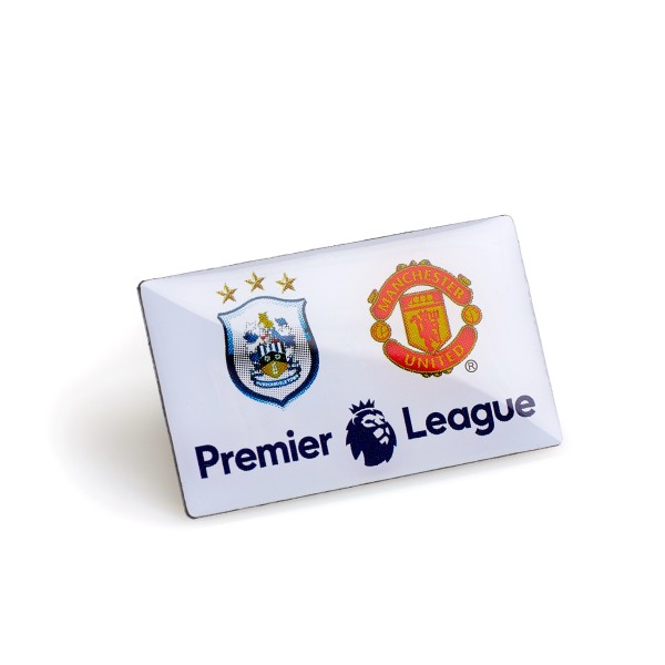 Rectangular printed pin badge with the Premier League logo and the team crests of Huddersfield Town and Manchester United to commemorate their league game.